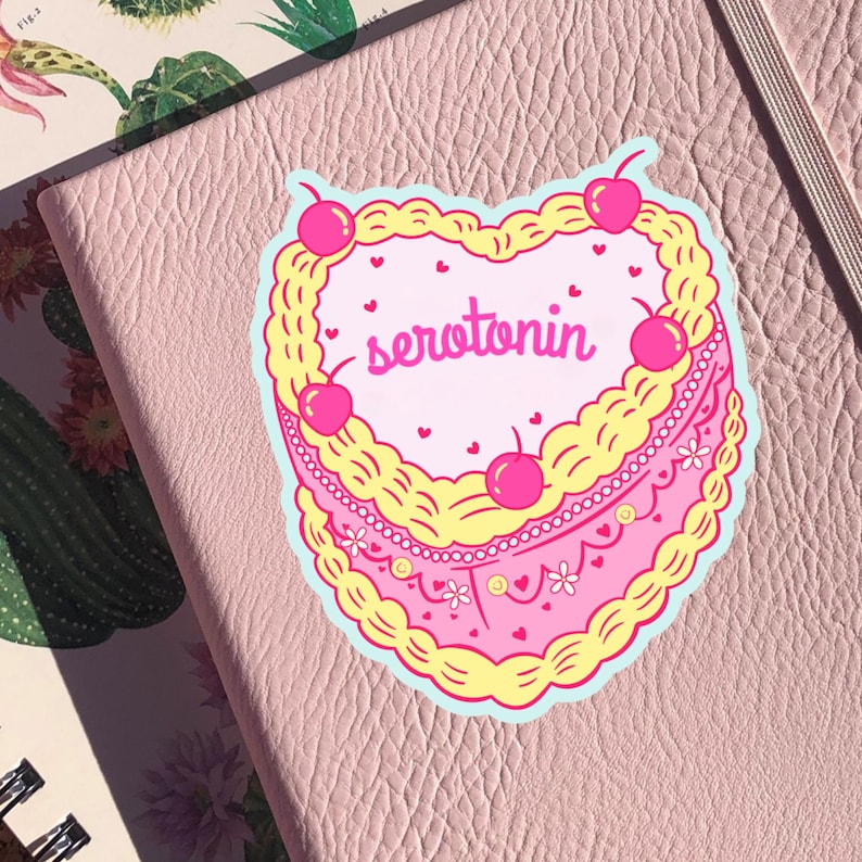May include: A pink and yellow heart-shaped cake with white frosting and cherries. The cake says "serotonin" in pink letters.