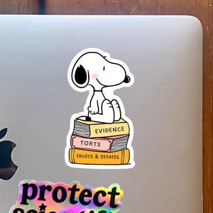 May include: A white Snoopy sticker sitting on a stack of books labeled "Evidence", "Torts", and "Trusts & Estates". The sticker is on a silver laptop. A holographic sticker that says "protect science" is at the bottom.