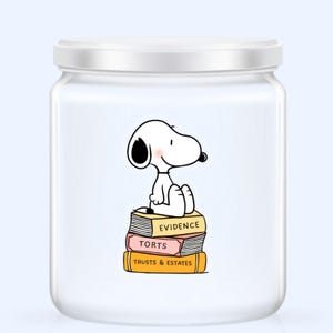 May include: A white glass jar with a white lid featuring a cartoon Snoopy character sitting on a stack of books. The books are labeled "Evidence", "Torts", and "Trusts & Estates". The jar has a light blue background.