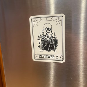 May include: A black and white illustration of a skeleton reading a book. The illustration is on a white card with black text that reads "REVIEWER 2" and "II".