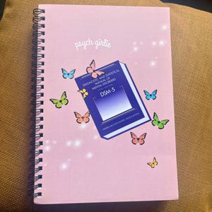 Puede incluir: Un cuaderno rosa con espiral con el texto "psych girlie" en cursiva. La portada presenta un gr&aacute;fico del libro DSM-5, mariposas y el texto "DIAGNOSTIC AND STATISTICAL MANUAL OF MENTAL DISORDERS". El cuaderno est&aacute; decorado con mariposas coloridas.