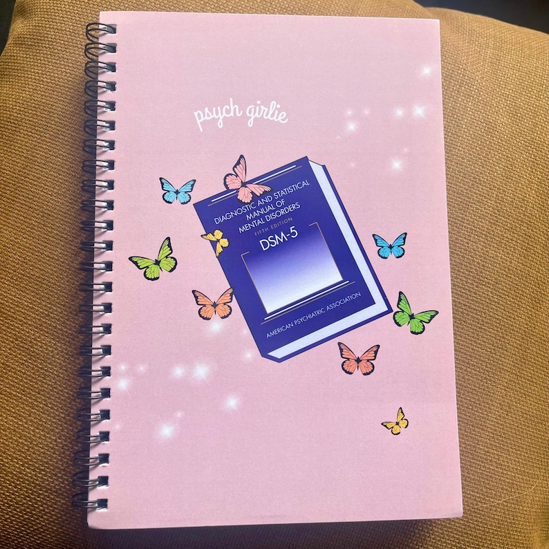 Puede incluir: Un cuaderno rosa con espiral con el texto "psych girlie" en cursiva. La portada presenta un gr&aacute;fico del libro DSM-5, mariposas y el texto "DIAGNOSTIC AND STATISTICAL MANUAL OF MENTAL DISORDERS". El cuaderno est&aacute; decorado con mariposas coloridas.