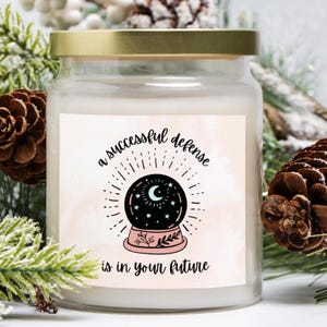May include: A white candle with a gold lid and a pink label that says "a successful defense is in your future". The label features a black and white illustration of a snow globe with a crescent moon and stars inside.