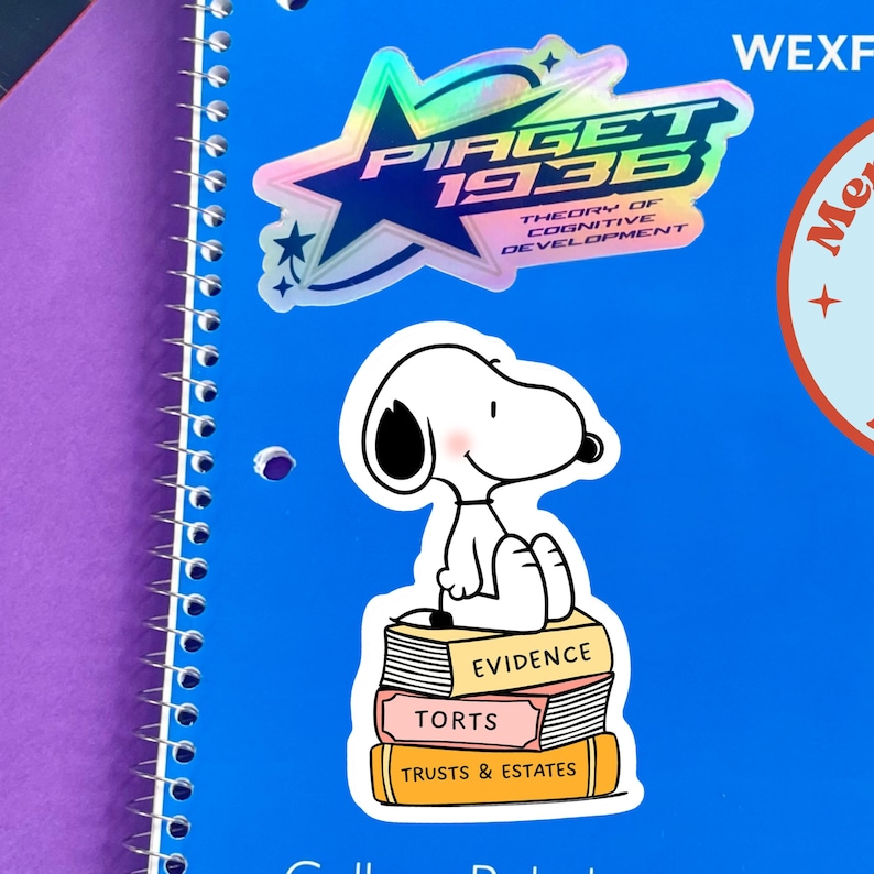 May include: A blue spiral notebook with stickers. One sticker features Snoopy sitting on books labeled "Evidence", "Torts", and "Trusts & Estates". Another sticker says "Piaget 1936 Theory of Cognitive Development".