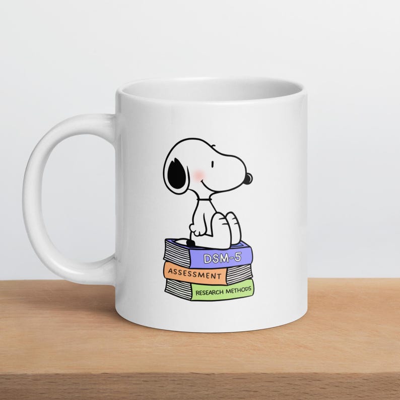 May include: White ceramic mug featuring a cartoon Snoopy character sitting on a stack of books. The books are labeled "DSM-5", "Assessment", and "Research Methods". The mug has a curved handle and is set on a wooden surface.