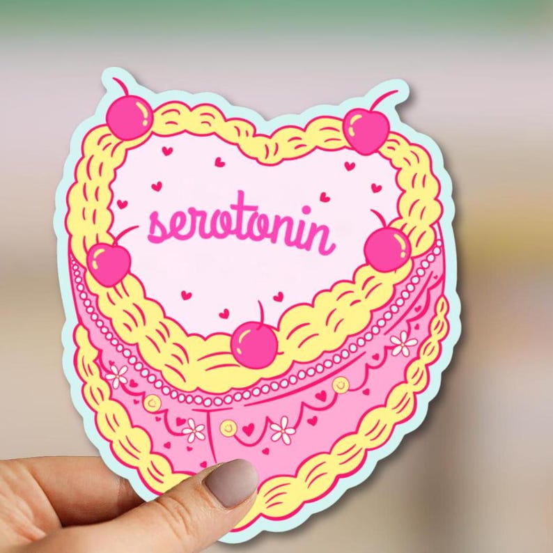 May include: A pink heart-shaped cake with yellow frosting and cherries on top. The cake says "serotonin" in pink letters.