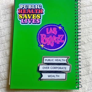 Pu&ograve; includere: Un quaderno verde con tre adesivi. L'adesivo superiore &egrave; un adesivo arcobaleno con il testo "Public Health Saves Lives". L'adesivo centrale &egrave; un adesivo glitterato viola e rosa con il testo "Lab Bratz". L'adesivo inferiore &egrave; un adesivo bianco con testo nero che dice "Public Health Over Corporate Wealth".