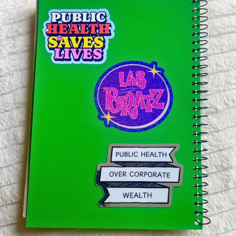 Pu&ograve; includere: Un quaderno verde con tre adesivi. L'adesivo superiore &egrave; un adesivo arcobaleno con il testo "Public Health Saves Lives". L'adesivo centrale &egrave; un adesivo glitterato viola e rosa con il testo "Lab Bratz". L'adesivo inferiore &egrave; un adesivo bianco con testo nero che dice "Public Health Over Corporate Wealth".
