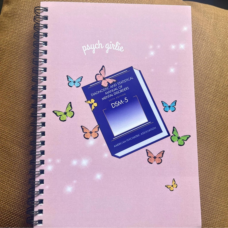 Puede incluir: Un cuaderno con espiral rosa con el texto "psych girlie" e ilustraciones de mariposas de colores. Un libro azul con el t&iacute;tulo "Diagnostic and Statistical Manual of Mental Disorders DSM-5" tambi&eacute;n est&aacute; presente.