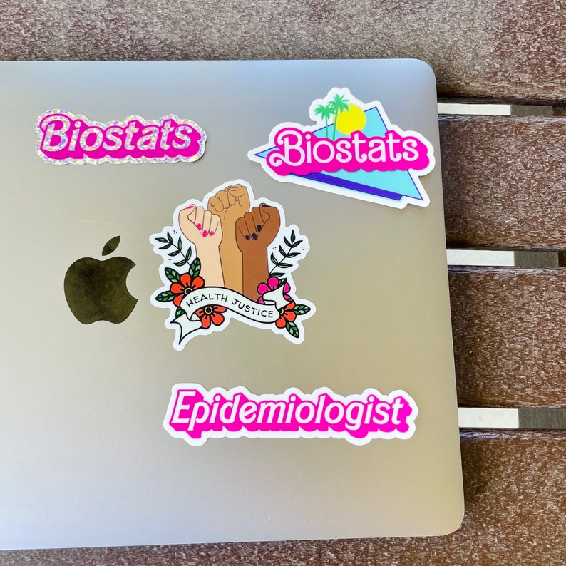 Pode incluir: Um laptop prateado com quatro adesivos. Os adesivos s&atilde;o rosa e apresentam as palavras "Biostats", "Epidemiologista" e "Justi&ccedil;a na sa&uacute;de". O adesivo de Justi&ccedil;a na sa&uacute;de apresenta um desenho de tr&ecirc;s punhos levantados com flores.