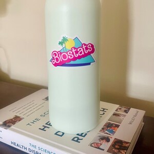 Pode incluir: Uma garrafa de &aacute;gua verde claro com um adesivo que diz "Biostats" em letras cor-de-rosa. O adesivo tem um design retro com uma palmeira, um sol amarelo e um fundo azul e branco.