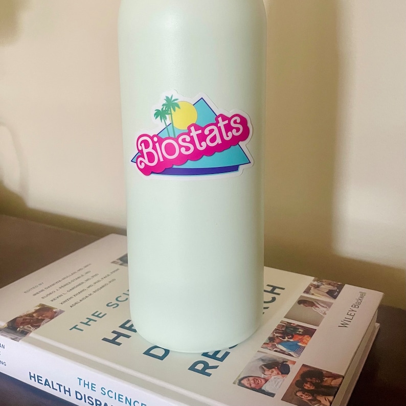 Pode incluir: Uma garrafa de &aacute;gua verde claro com um adesivo que diz "Biostats" em letras cor-de-rosa. O adesivo tem um design retro com uma palmeira, um sol amarelo e um fundo azul e branco.