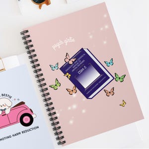 May include: A spiral-bound notebook with a pink cover featuring the text "psych girlie" and butterfly illustrations. A graphic of the DSM-5 book is also on the cover. The notebook is open to reveal the spiral binding.