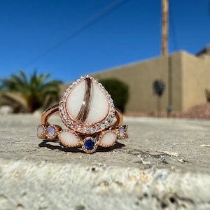 Può includere: Due anelli d'oro con pietre bianche e una pietra blu. L'anello superiore presenta una pietra a forma di lacrima con un motivo a spirale marrone.