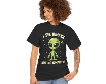 May include: Black t-shirt featuring a green alien graphic with large black eyes. The text "I SEE HUMANS BUT NO HUMANITY!" is printed above and below the alien illustration. The design includes stars and a crescent moon.