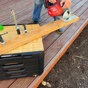 May include: A person uses a red circular saw to cut a piece of wood on a black workbench with drawers. The workbench has a ruler and markings on the surface. There are two green and silver clamps on the workbench.