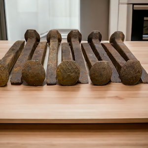 May include: Eight rusty railroad spikes laid out on a light brown wooden surface. The spikes are all the same size and shape, with a square head and a pointed tip. They are all slightly bent and have a rough, textured surface.