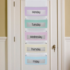May include: A white over-the-door organizer with five pastel-colored pockets labeled with the days of the week: Monday, Tuesday, Wednesday, Thursday, and Friday. The organizer hangs from two hooks.