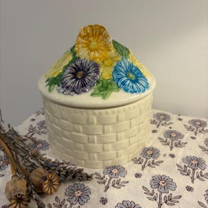 May include: A white ceramic jar with a brick pattern and a lid decorated with colourful flowers. The flowers are yellow, blue, purple, and green.