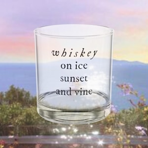 Puede incluir: Un vaso de whisky transparente con las palabras "whiskey on ice sunset and vine" impresas en escritura negra. El vaso está vacío y sobre un fondo borroso de una puesta de sol sobre el agua.