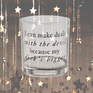 May include: A clear glass tumbler with the phrase "I can make deals with the devil because my dick's bigger" printed in black. The background features hanging gold stars.
