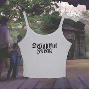 Puede incluir: Una camiseta sin mangas gris claro con tirantes finos. El texto "Delightful Freak" está impreso en negro, con letras góticas en la parte delantera. El fondo presenta una escena borrosa al aire libre con árboles y una estructura de piedra.