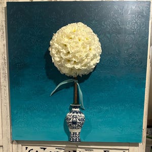 May include: A decorative floral arrangement featuring a large, spherical, cream-colored flower atop a slender brown stem. The stem is secured in a blue and white patterned vase. The backdrop is a square canvas with a teal ombre effect and a subtle damask pattern.