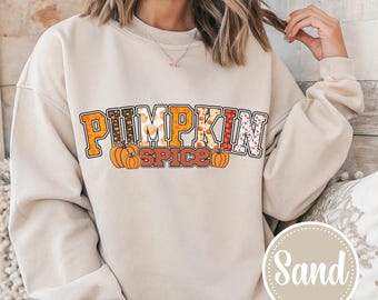 May include: A sand-colored crewneck sweatshirt featuring the text PUMPKIN SPICE in a collegiate font. The letters are filled with various autumn-themed patterns including pumpkins and leopard prints, with two orange pumpkin illustrations below.