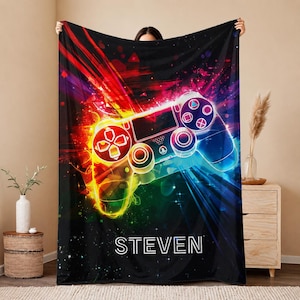 May include: A black fleece blanket featuring a vibrant, multicolored graphic of a video game controller surrounded by bright red, yellow, and blue light streaks. The name STEVEN is printed in white capital letters at the bottom of the design.