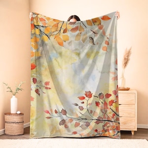 Op de afbeelding: Een rechthoekige plaid met een aquarelprint van boomtakken met herfstbladeren in tinten oranje, rood, geel en bruin tegen een zachte, gedempte achtergrond van lichtgeel, crème en lichtgrijze tinten.