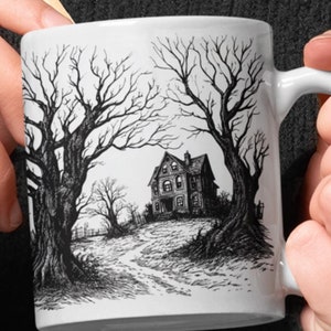 May include: White ceramic mug with a black and white illustration of a spooky house with bare trees on either side of a path leading to the house.