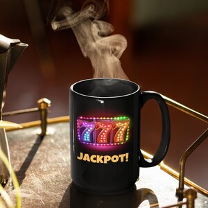May include: Black coffee mug with a colorful graphic of three sevens and the word "JACKPOT!" in yellow. Steam rises from the mug, suggesting a hot beverage. The mug is on a table with a gold-colored metal frame.