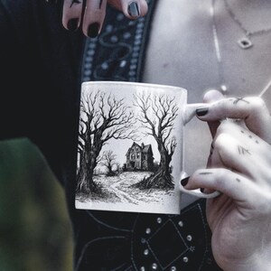 May include: White ceramic mug with a black and white illustration of a spooky house with trees on a path. The house has a pointed roof and windows.