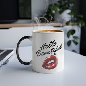 May include: White ceramic mug with a black handle. The mug has a black and white graphic that says "Hello Beautiful" with a red lipstick print underneath.