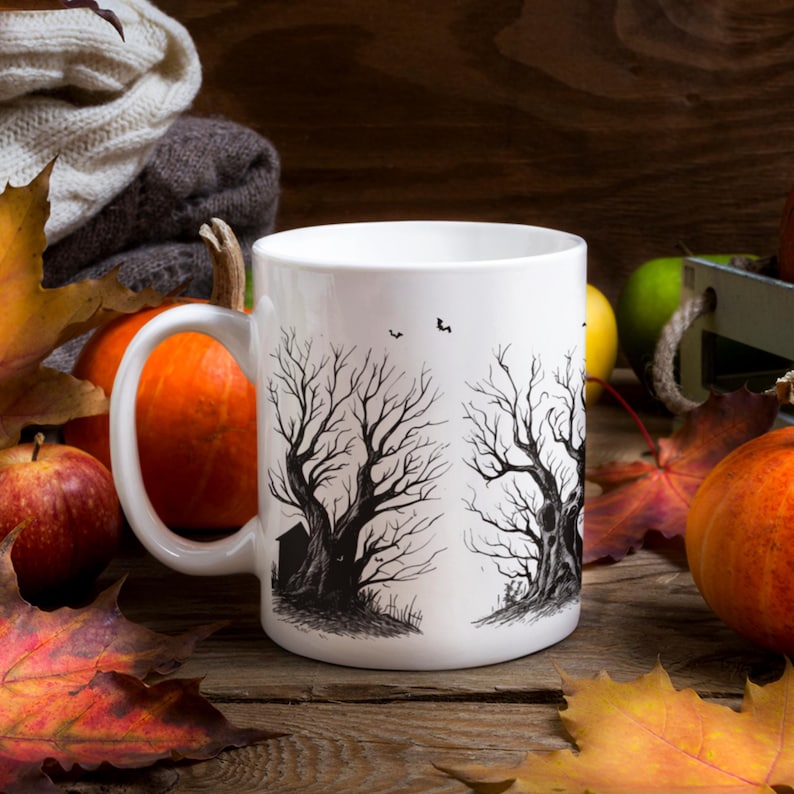 May include: White ceramic mug with a black and white illustration of two bare trees with a small house in the background. The mug is surrounded by fall leaves and pumpkins.