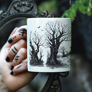 May include: White ceramic mug with a black and white illustration of two bare trees with branches reaching towards the sky. The trees are in a forest setting with a path leading through the trees.