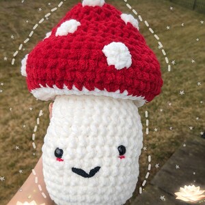 May include: A crocheted mushroom plush toy with a red cap and white spots. The mushroom has a white stem with a smiling face and black eyes.