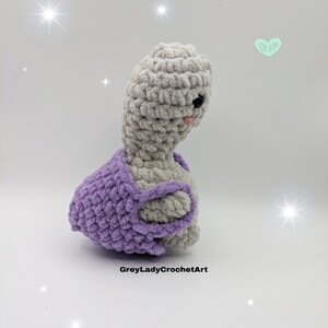 May include: A crocheted gray and purple turtle with a sweet expression. The turtle is wearing a purple crocheted shell.