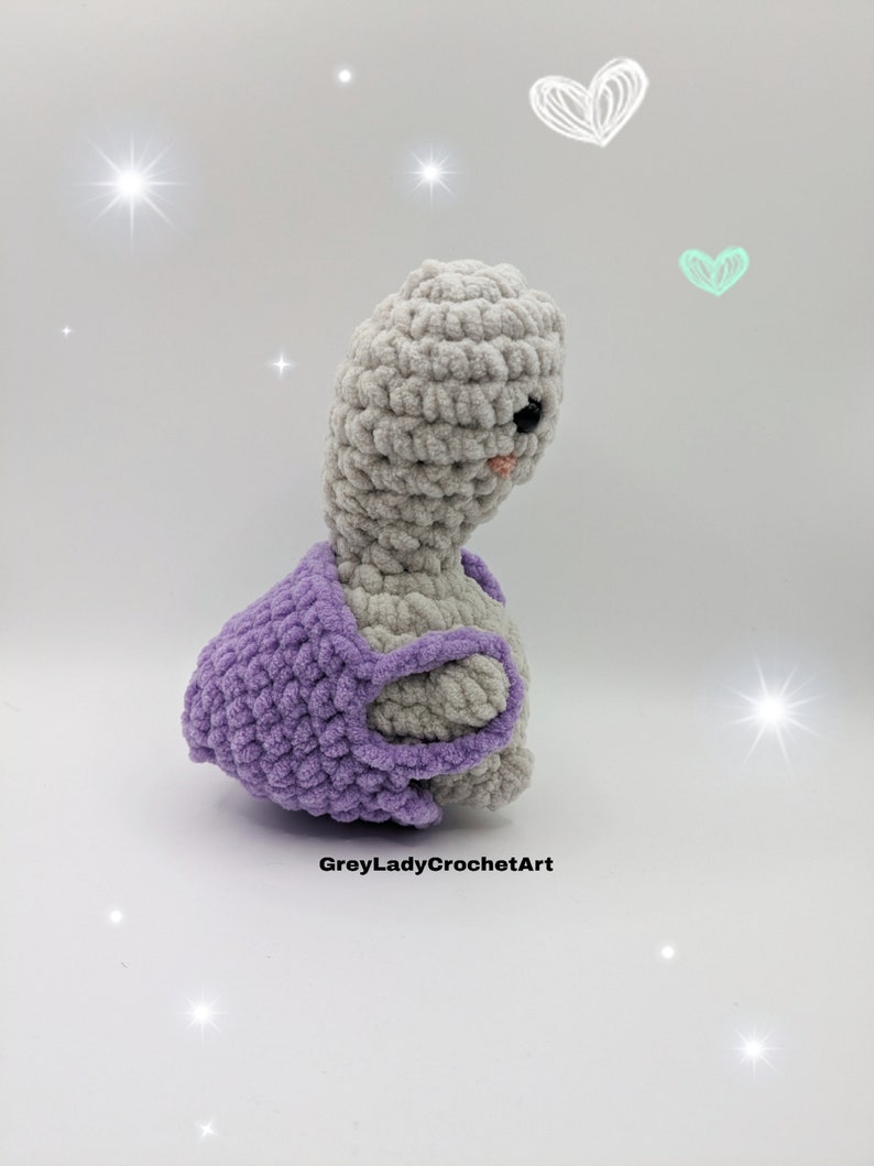 May include: A crocheted grey and purple turtle with a sweet expression. The turtle is wearing a purple crocheted shell.