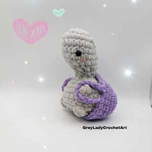 May include: A crocheted grey and purple plush toy in the shape of a turtle. The turtle has a small pink nose and black eyes.