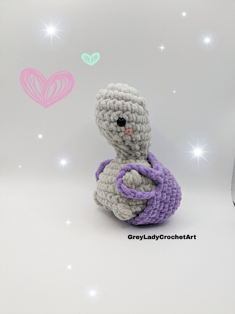 May include: A crocheted grey and purple plush toy in the shape of a turtle. The turtle has a small pink nose and black eyes.