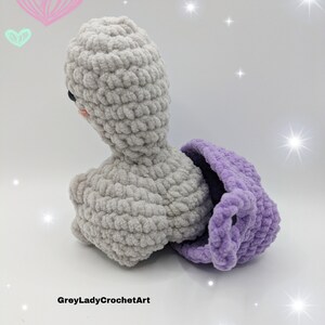 May include: A crocheted grey and purple stuffed animal with a heart-shaped head and a purple shell. The animal is sitting on a white surface with a light pink heart and a light green heart in the background.