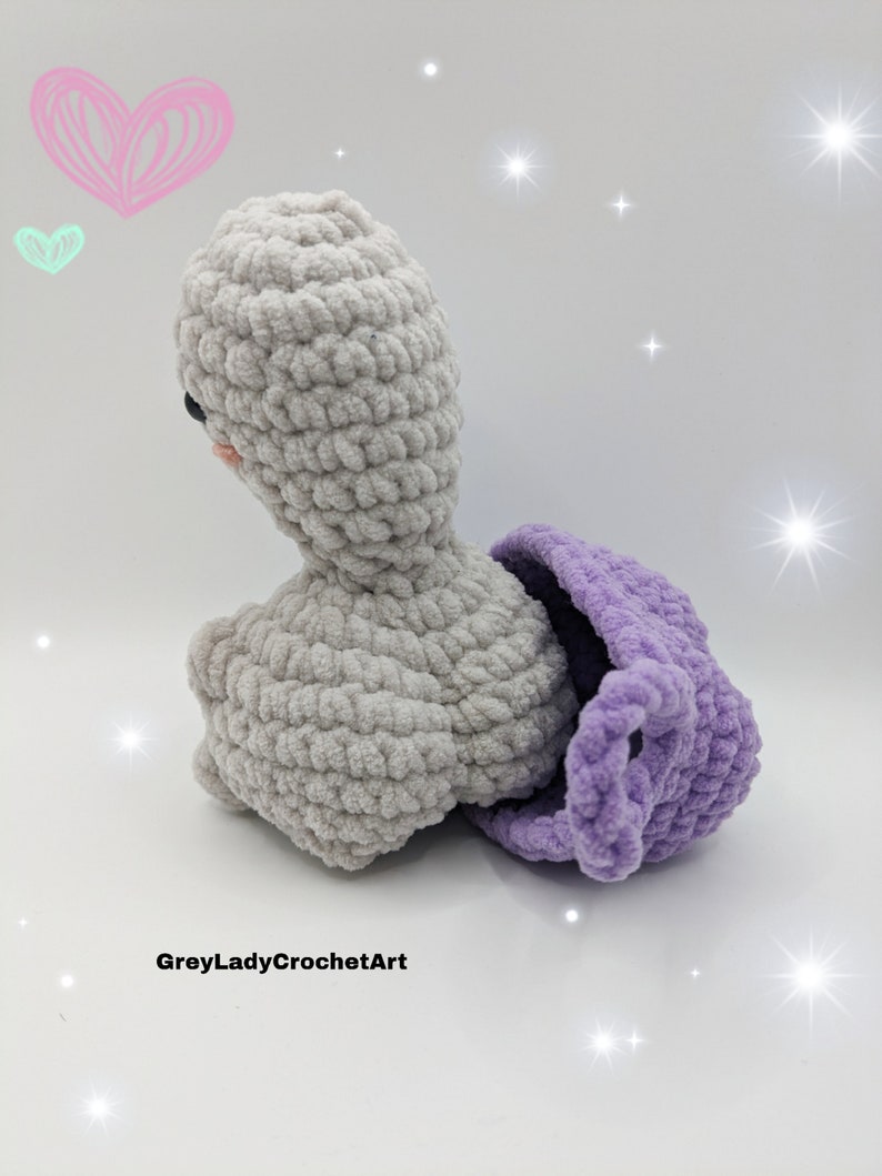May include: A crocheted grey and purple stuffed animal with a heart-shaped head and a purple shell. The animal is sitting on a white surface with a light pink heart and a light green heart in the background.