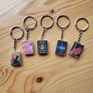 May include: Six miniature book keychains featuring covers from popular young adult novels. The keychains are silver and feature the following titles: "Harry Potter", "The Apprentice to the Villain", "What the River Knows", "Caraval", and "Red Rising".