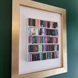May include: A framed artwork featuring a miniature bookshelf filled with colorful books. The bookshelf is white, set against a white background, and framed in gold. The books have various colors and sizes, creating a vibrant display.