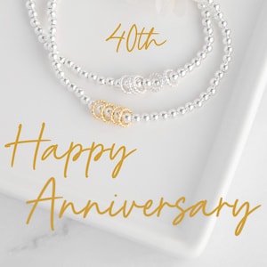 May include: A silver beaded bracelet with a gold charm of interlocking rings. The bracelet is on a white surface with a white flower and the text "40th Happy Anniversary".