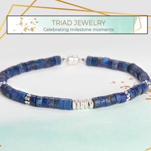 May include: A blue beaded bracelet with silver accents and a magnetic clasp. The bracelet is made of small, square-shaped beads. The text "TRIAD JEWELRY Celebrating milestone moments" is in the background.