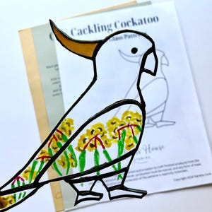 May include: A white cockatoo stained glass pattern with a yellow beak and black outline. The cockatoo is decorated with yellow flowers and green leaves. The pattern is labeled "Cackling Cockatoo".