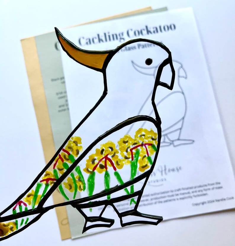 May include: A white cockatoo stained glass pattern with a yellow beak and black outline. The cockatoo is decorated with yellow flowers and green leaves. The pattern is labeled "Cackling Cockatoo".