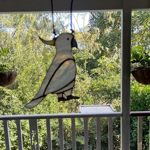 May include: A white stained glass cockatoo bird hanging from a porch ceiling. The bird is suspended in front of a window with a view of green trees.